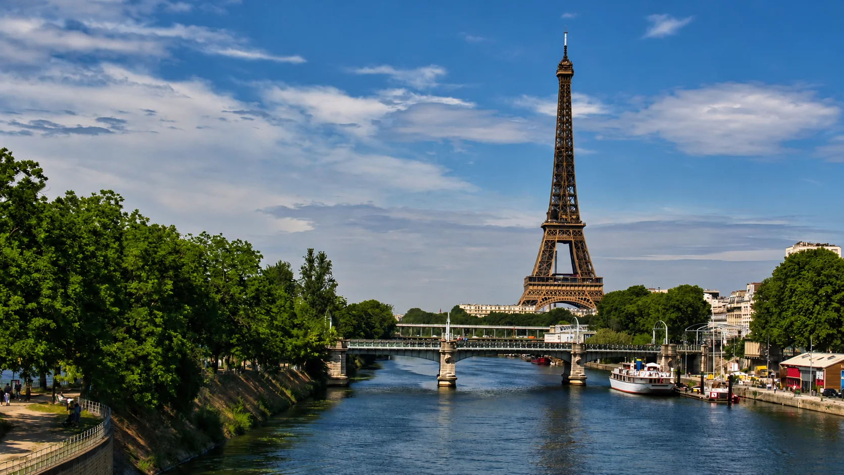 The Eiffel Tower beside the Seine in Paris, with a bridge, boats and tree-lined banks in the foreground