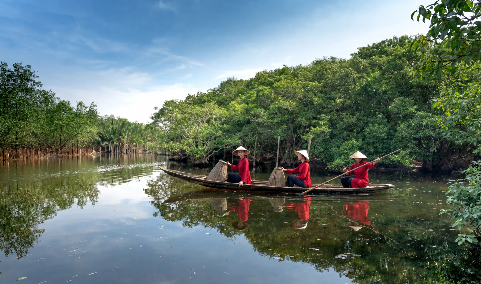Kolme naista punaisissa áo dài -asuissa ja perinteisissä kartiohatuissa puuveneessä mangrovepuiden reunustamalla joella Vietnamissa